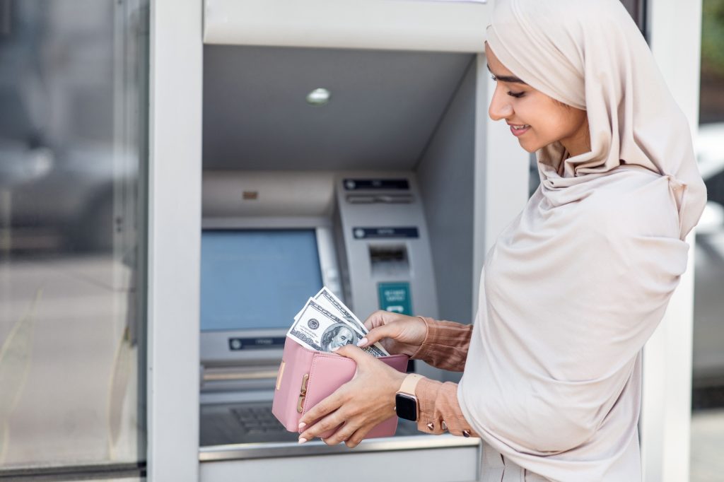 Satisfied cute millennial muslim woman in hijab puts many dollars in wallet near ATM outdoor, empty